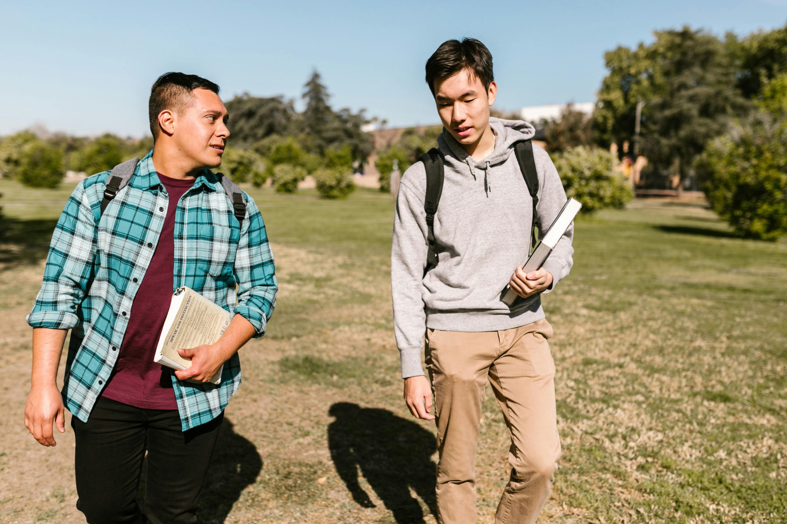 Two male college students walk on a sunny campus day holding textbooks and backpacks.