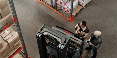 Two workers in a warehouse discussing logistics near a forklift captured from above.